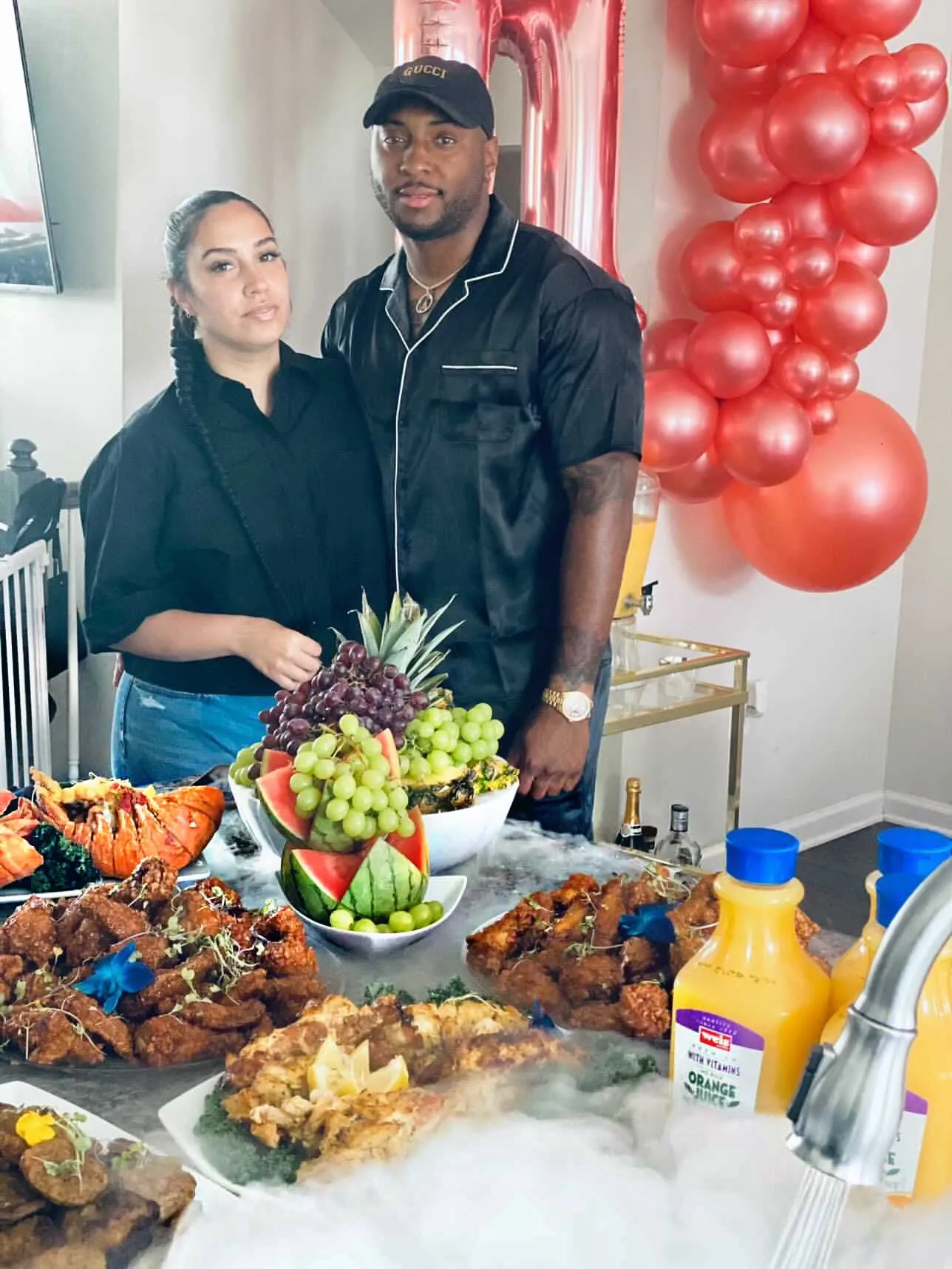 Two people in black standing behind a buffet of wings, lobster tails, a fruit bowl and orange juice, with red balloons behind them