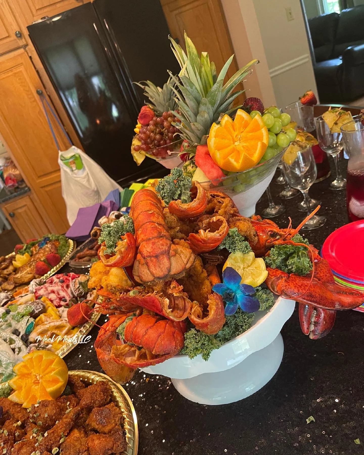 Seafood tower of lobster, crab and kale on a white pedestal stand, with a carved fruit centerpiece and wine glasses behind it
