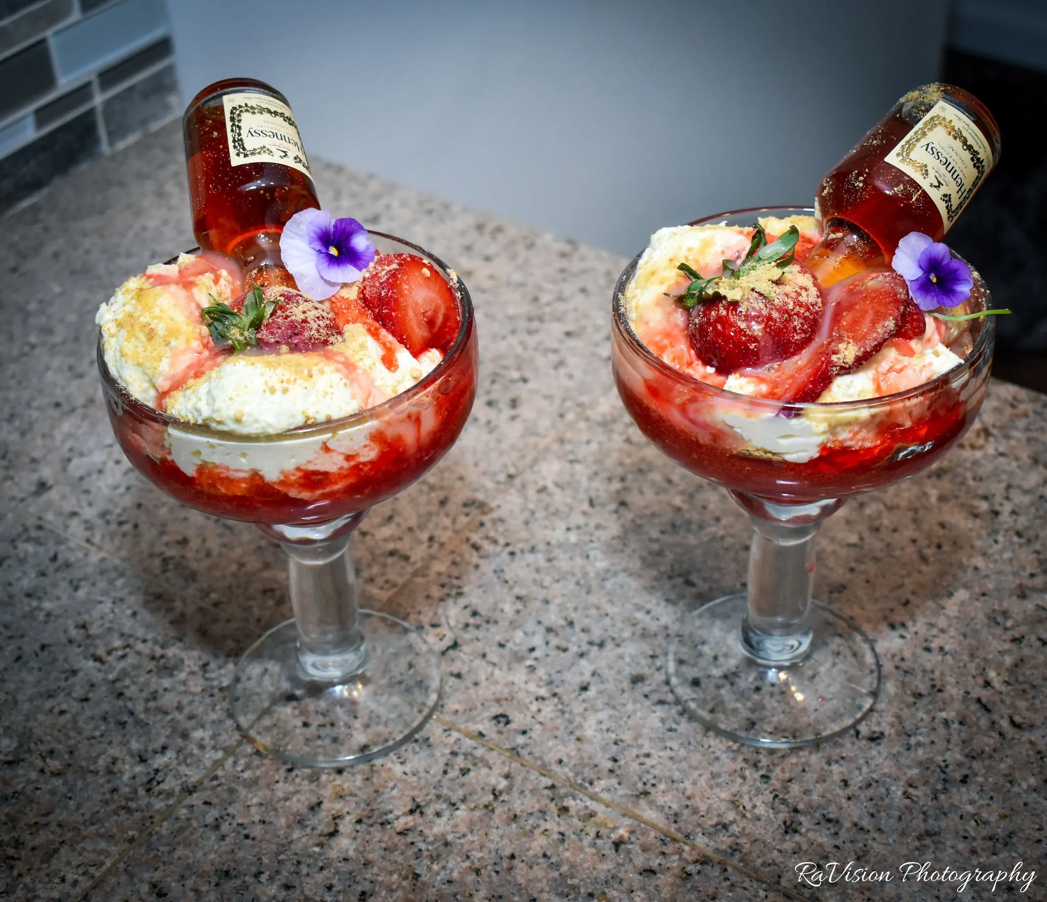 Two Strawberry Hennessy Cheesecake desserts in stemmed glasses, each topped with a mini Hennessy bottle and a purple flower