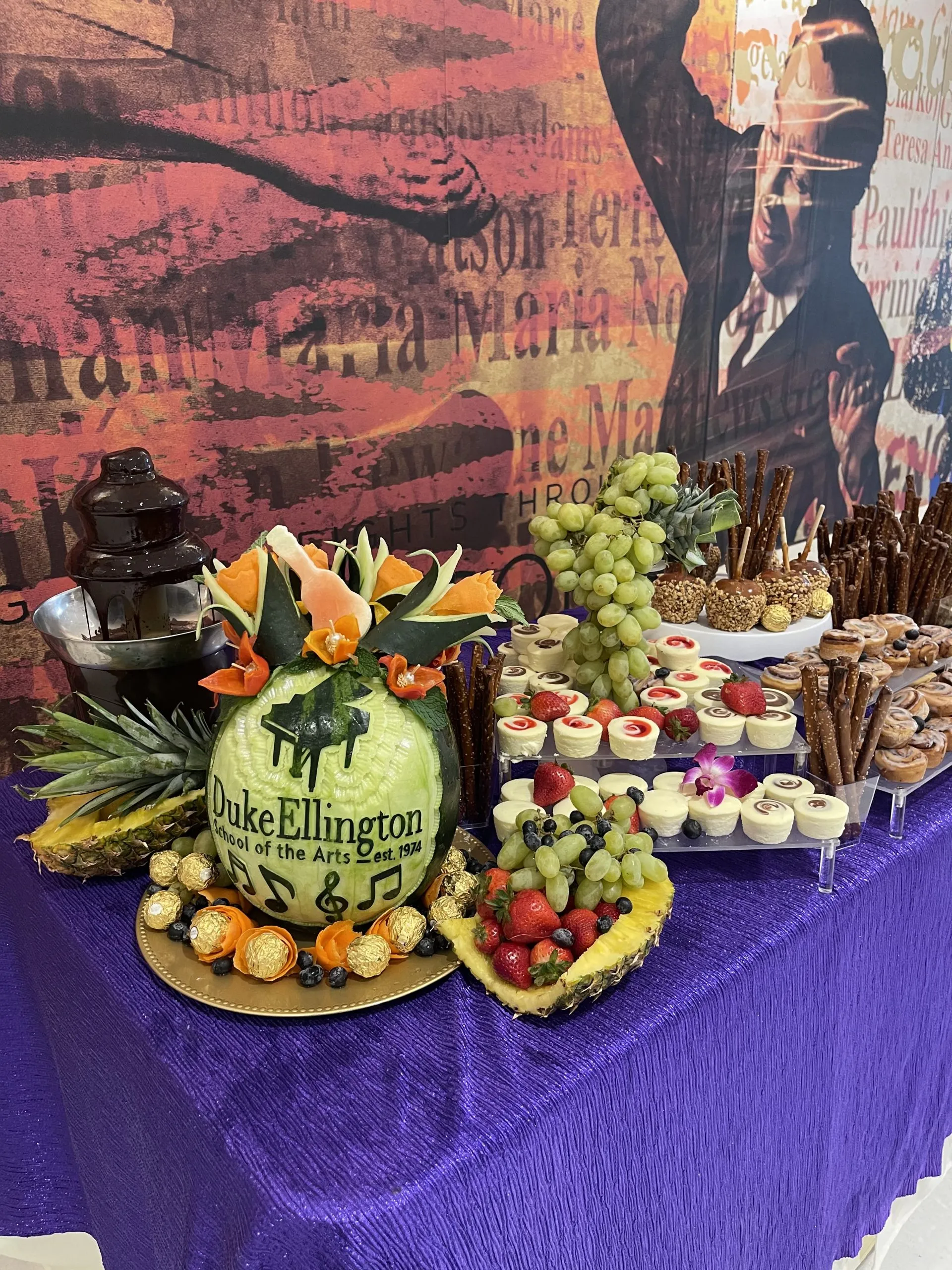 Dessert and fruit table with a carved melon reading Duke Ellington School of the Arts, a chocolate fountain and chocolate-dipped pretzel rods