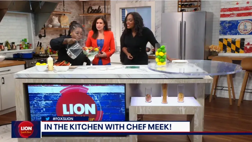 Chef Meek pours from a pitcher into a stemmed glass on the FOX 5 LION Lunch Hour kitchen set beside two hosts, under the on-screen banner In the Kitchen with Chef Meek